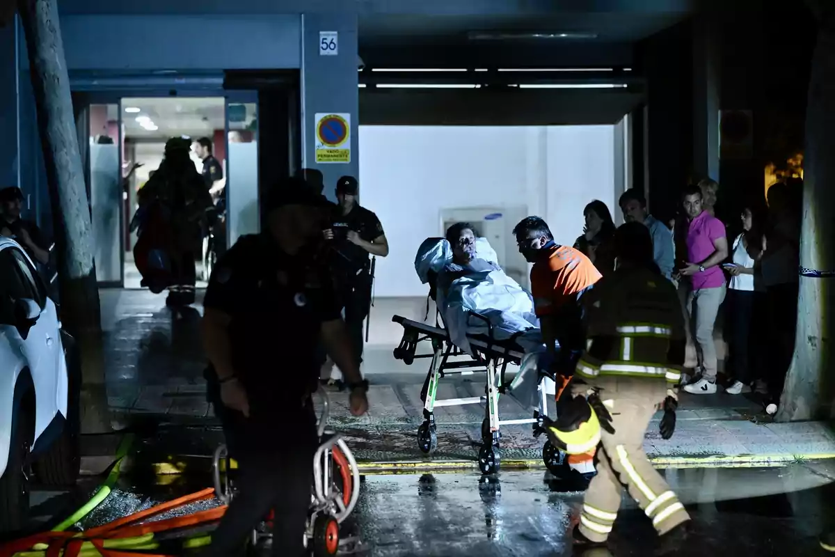 Personal de emergencia traslada a una persona en camilla fuera de un edificio durante la noche mientras varias personas observan la escena Personal de emergencia traslada a una persona en camilla fuera de un edificio durante la noche mientras varias personas observan la escena