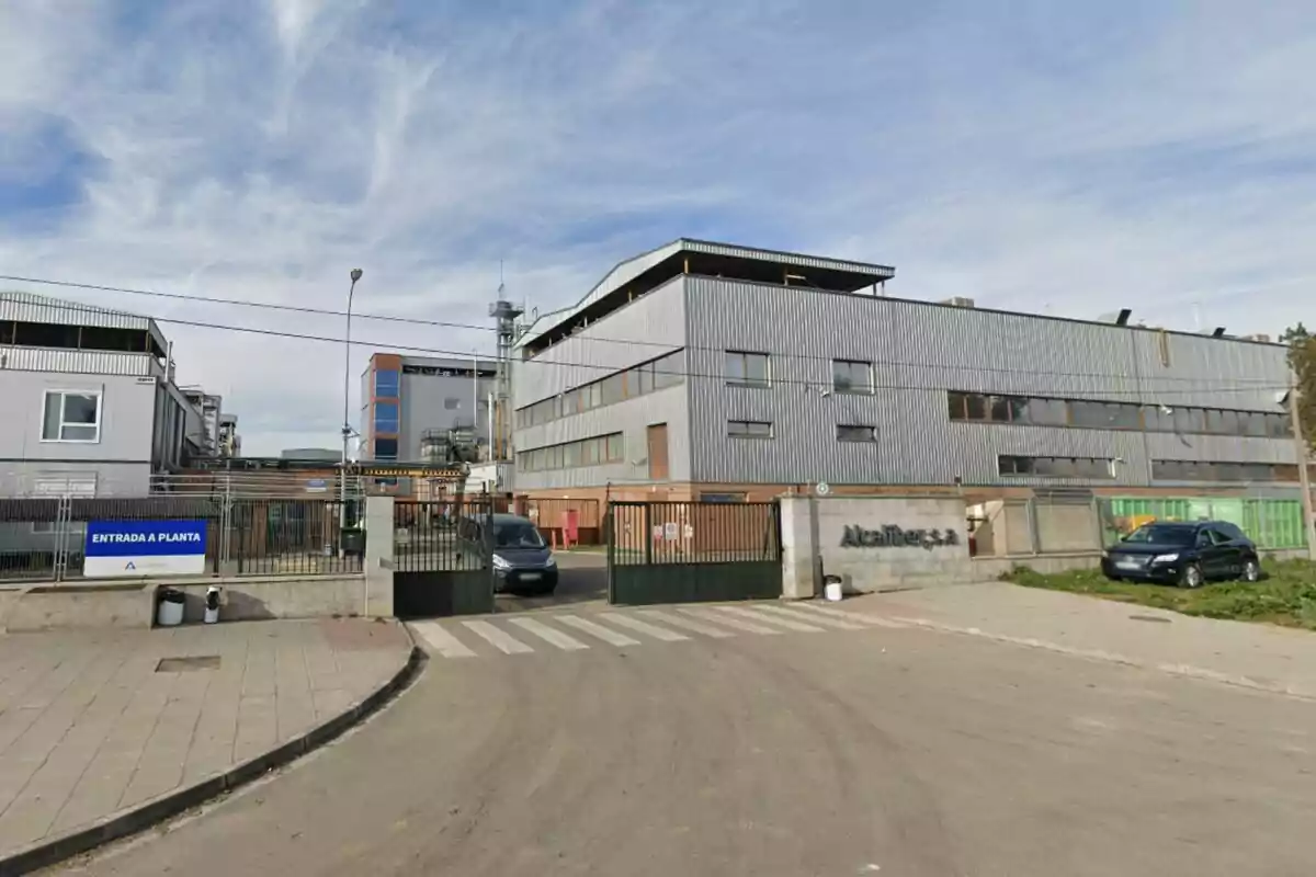 Edificio industrial con fachada de metal y ventanas rectangulares, rodeado de una cerca y con un cartel que indica "Entrada a Planta".