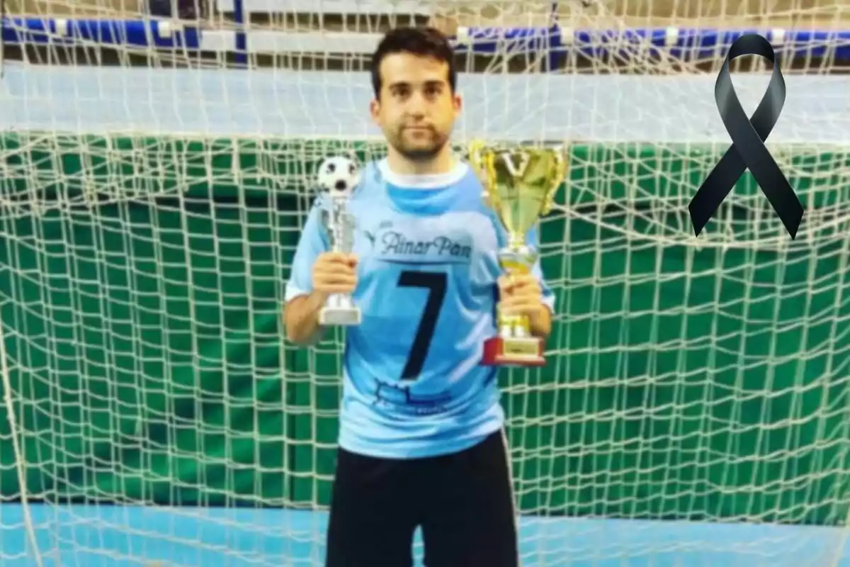 Un jugador de fútbol sala con camiseta azul y el número 7 sostiene dos trofeos frente a una portería, con un lazo negro en la esquina superior derecha. Un jugador de fútbol sala con camiseta azul y el número 7 sostiene dos trofeos frente a una portería, con un lazo negro en la esquina superior derecha.