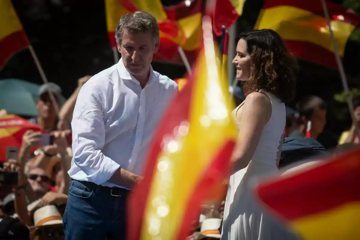 Dos personas conversan en un evento al aire libre rodeados de banderas de España y un grupo de personas al fondo