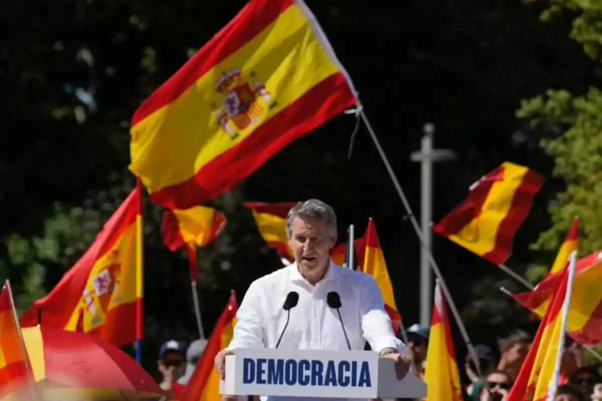 Hombre hablando en un mitin al aire libre rodeado de banderas de España y un cartel que dice democracia Hombre hablando en un mitin al aire libre rodeado de banderas de España y un cartel que dice democracia