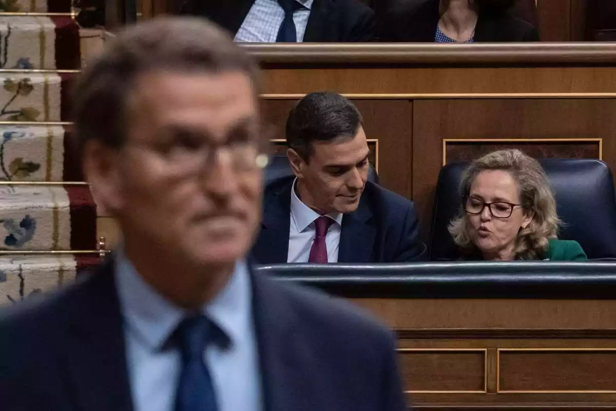 Alberto Núñez Feijóo en primer plano, borroso, y de fondo Pedro Sánchez y Nadia Calviño hablando sentados en sus escaños del Congreso de los Diputados