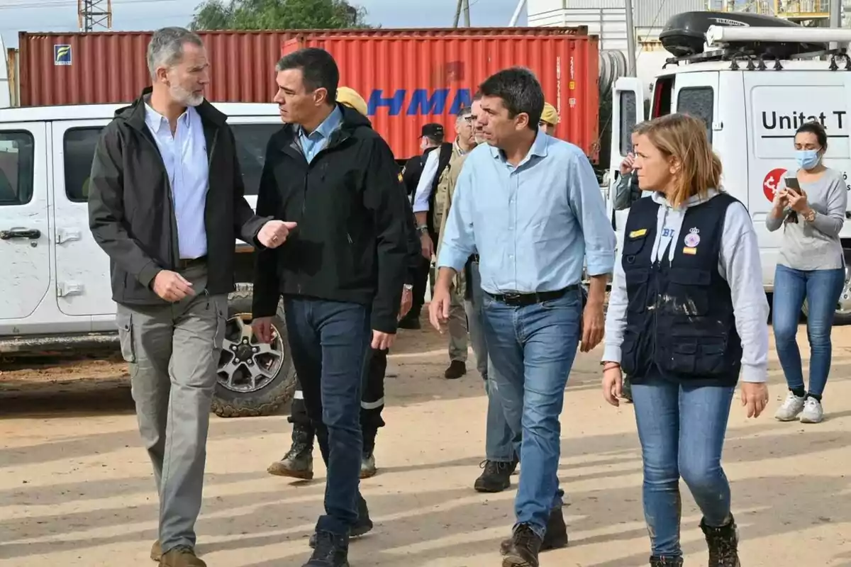 Felipe VI, Pedro Sánchez y Carles Mazón camina y conversa en un área al aire libre con vehículos y contenedores de fondo.