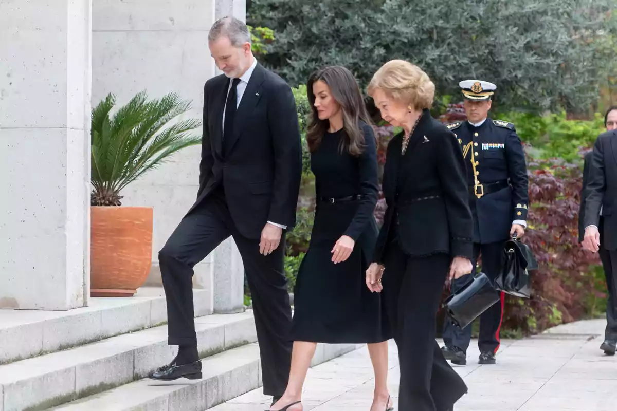 Felipe VI, la reina Letizia y la reina Sofía vestidas de negro suben unas escaleras en un entorno exterior acompañadas por un hombre con uniforme militar Felipe VI, la reina Letizia y la reina Sofía vestidas de negro suben unas escaleras en un entorno exterior acompañadas por un hombre con uniforme militar
