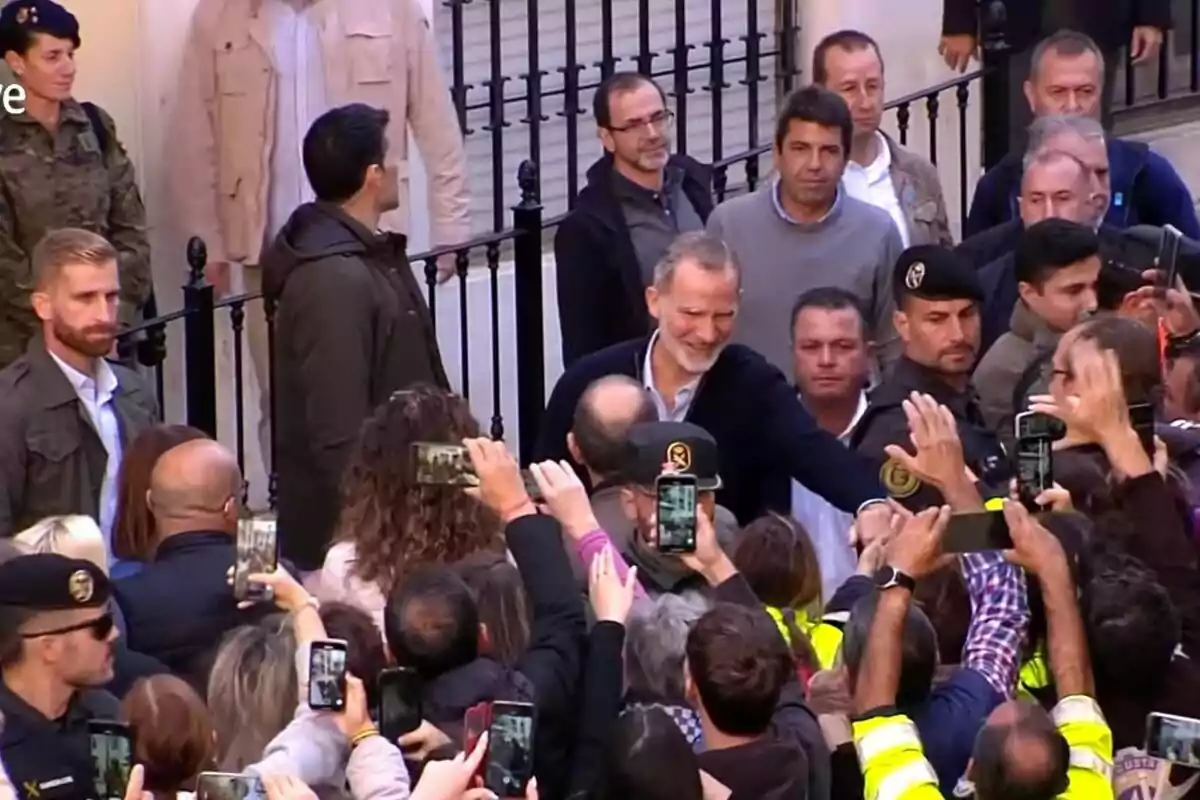 Felipe VI sonriente saluda a una multitud mientras varias personas toman fotos con sus teléfonos móviles.