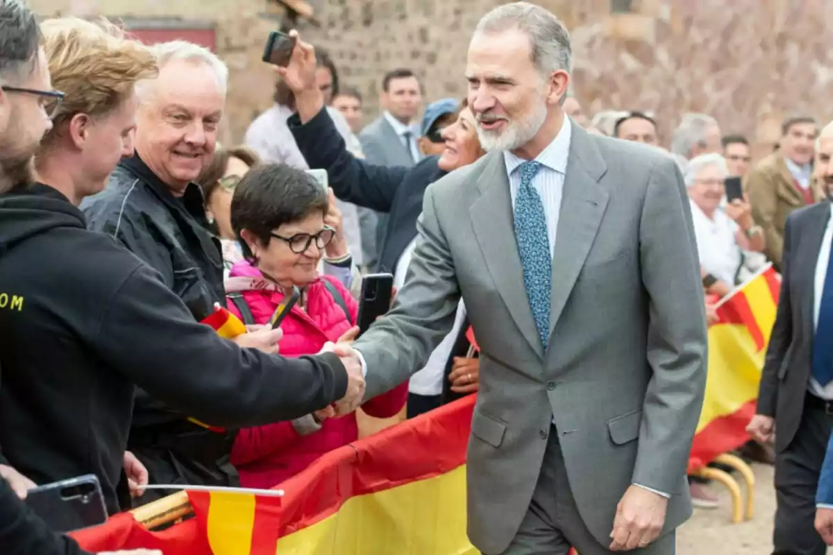 Felipe VI con traje gris saluda a personas detrás de una valla decorada con banderas de España mientras varios asistentes toman fotos y sonríen Felipe VI con traje gris saluda a personas detrás de una valla decorada con banderas de España mientras varios asistentes toman fotos y sonríen