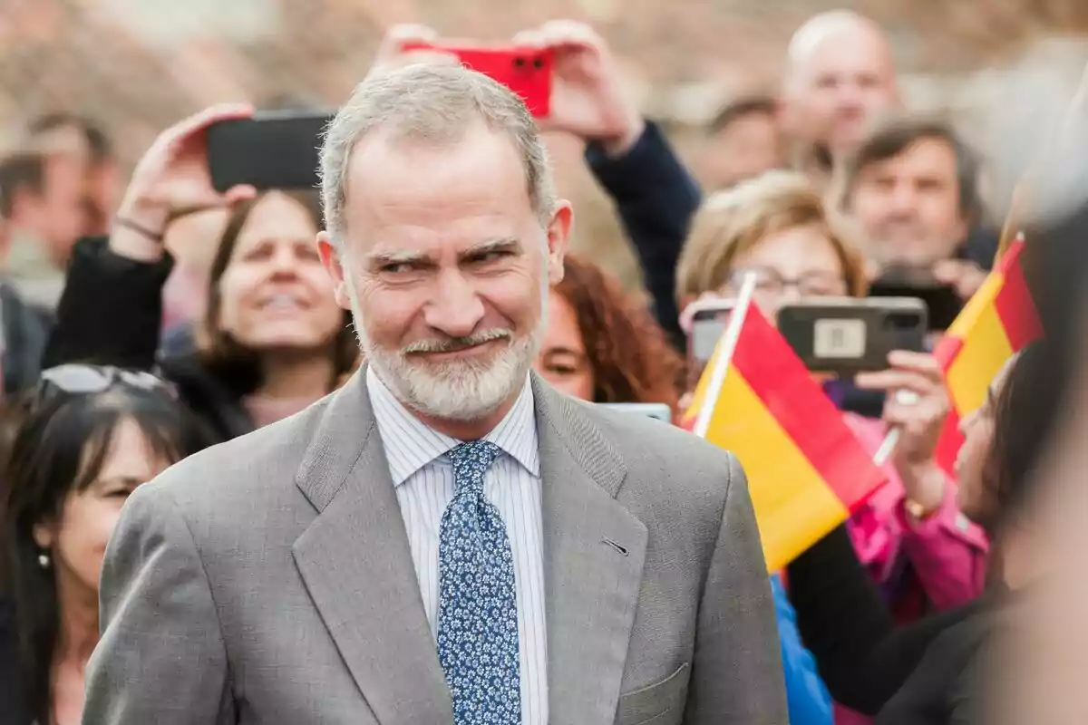 Felipe VI con traje gris y corbata azul sonríe mientras una multitud lo fotografía y ondea banderas de España Felipe VI con traje gris y corbata azul sonríe mientras una multitud lo fotografía y ondea banderas de España