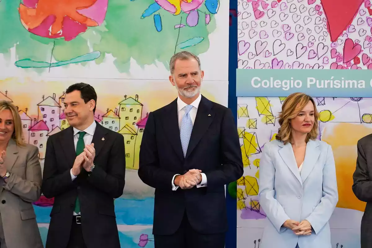 Varias personas vestidas de manera formal posan frente a un fondo colorido con dibujos infantiles y un cartel que dice Colegio Purísima Varias personas vestidas de manera formal posan frente a un fondo colorido con dibujos infantiles y un cartel que dice Colegio Purísima