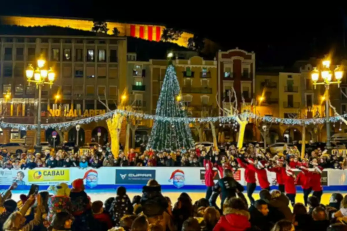 Una plaza iluminada con un árbol de Navidad y una pista de patinaje sobre hielo rodeada de personas observando un espectáculo.