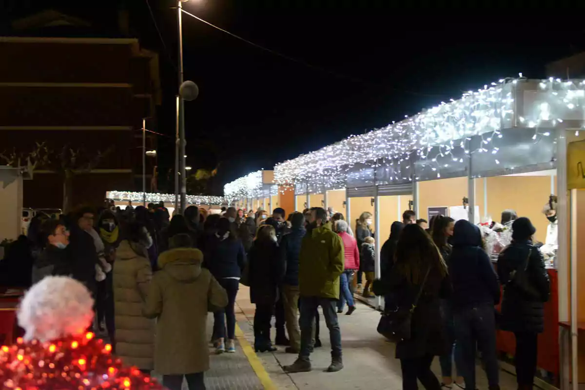 Una multitud de personas pasea por un mercado nocturno al aire libre con luces decorativas blancas en los puestos.