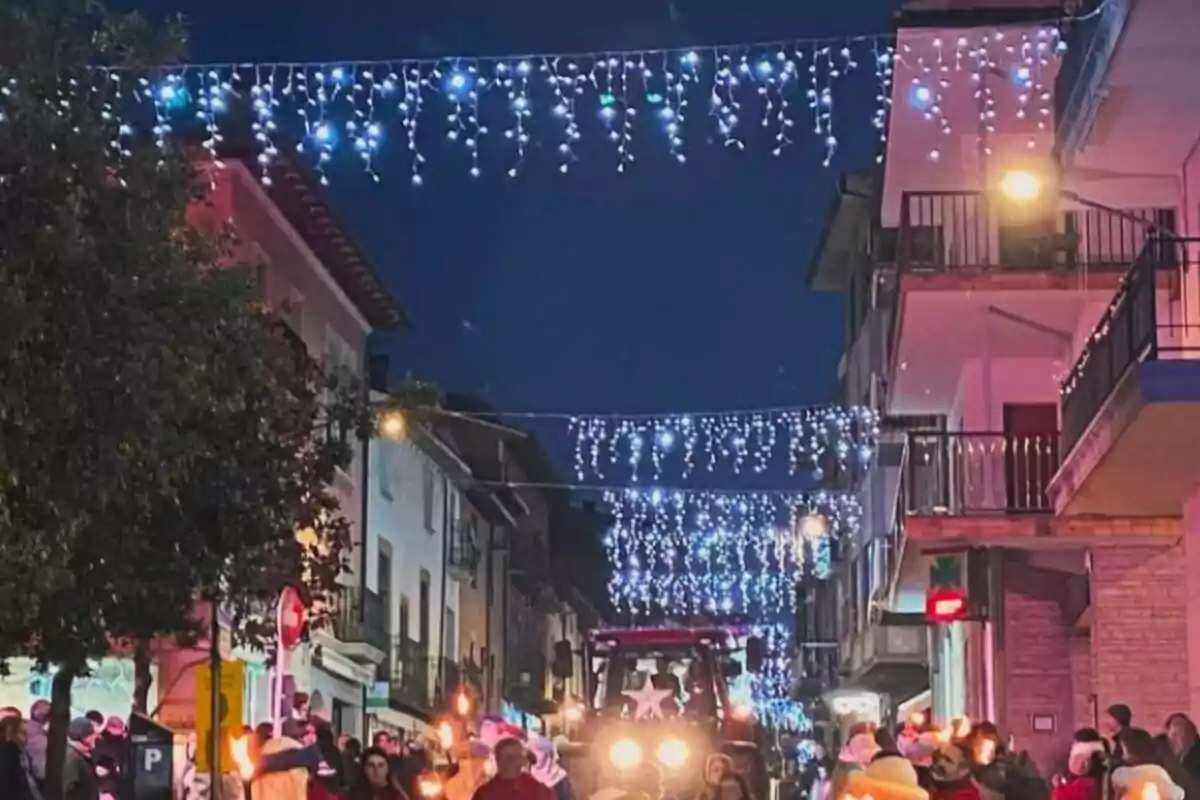 Una calle decorada con luces navideñas colgantes y un grupo de personas caminando con antorchas.