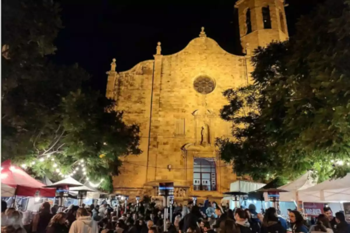 Una iglesia iluminada por la noche con un mercado al aire libre lleno de gente y puestos con luces.