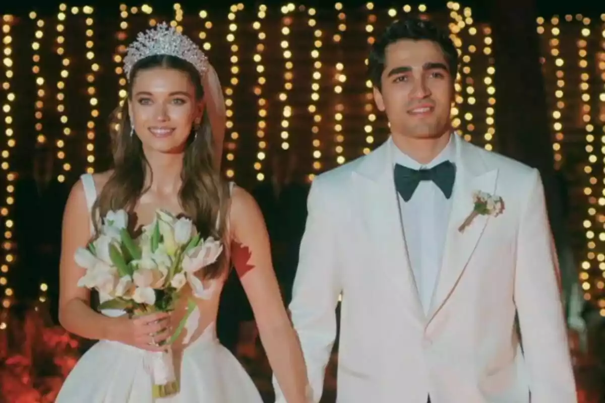 Una pareja formada por Ferit y Seyran vestida de novios sonríe mientras camina de la mano en un ambiente iluminado con luces brillantes en Una nueva vida. Una pareja formada por Ferit y Seyran vestida de novios sonríe mientras camina de la mano en un ambiente iluminado con luces brillantes en Una nueva vida.
