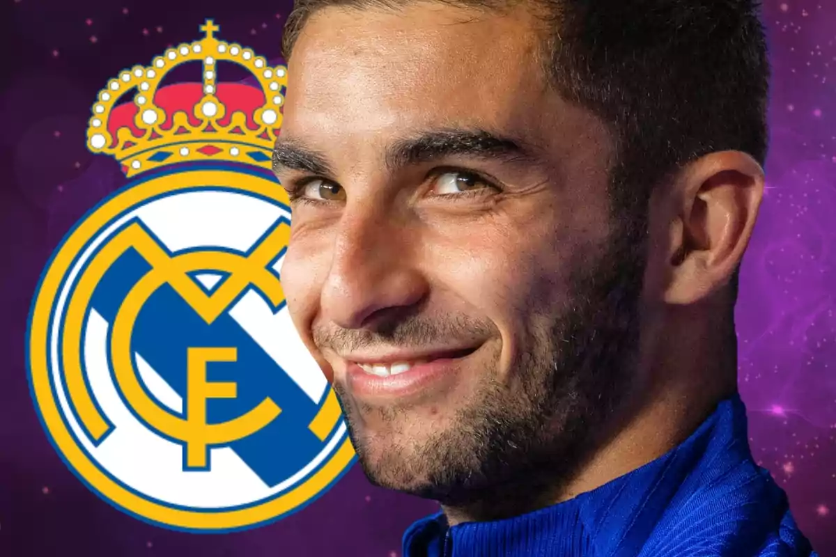 Un hombre sonriente con barba y cabello corto, vestido con una camiseta azul, aparece en primer plano con el escudo del Real Madrid de fondo.