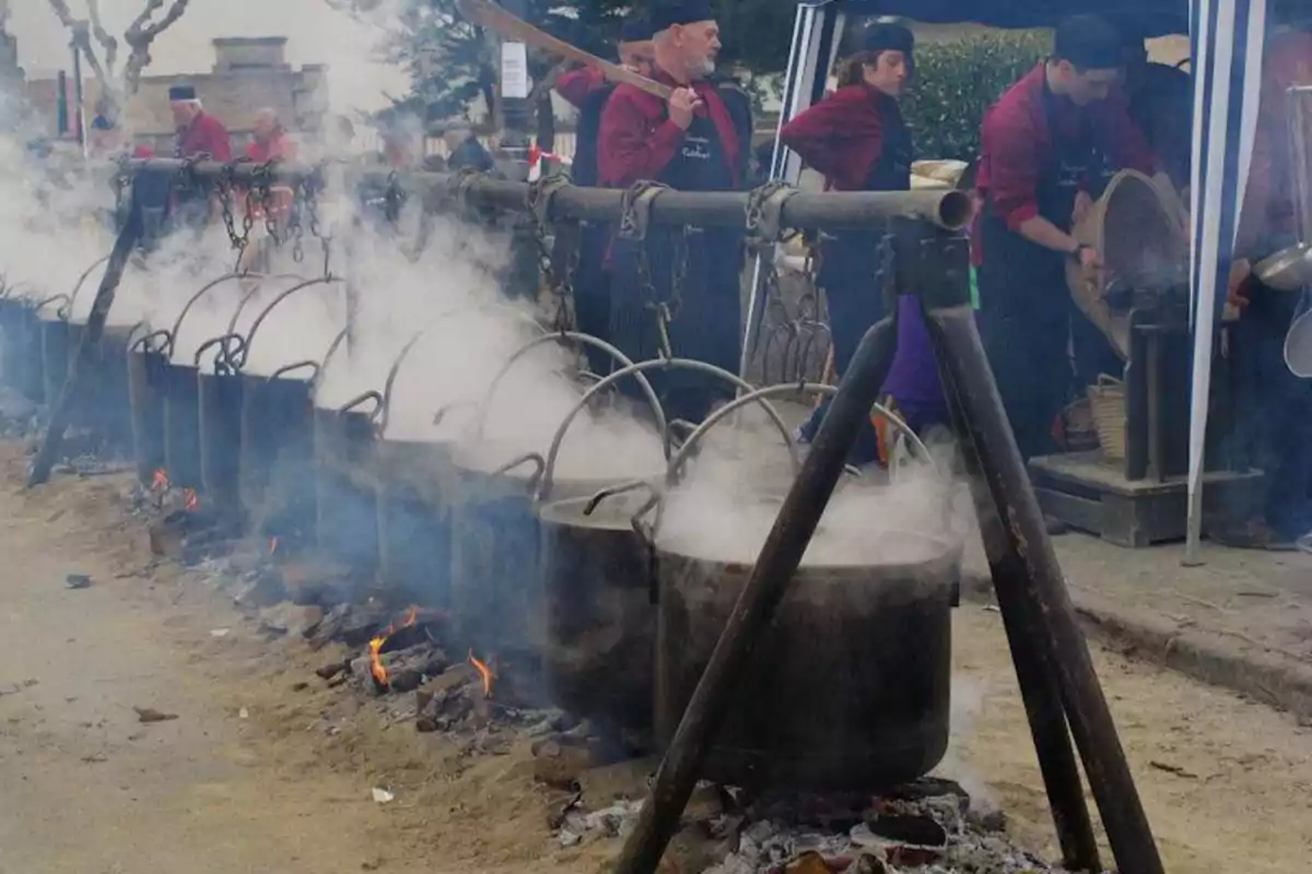 Personas cocinando al aire libre con grandes ollas colgantes sobre fuego de leña, rodeadas de humo.