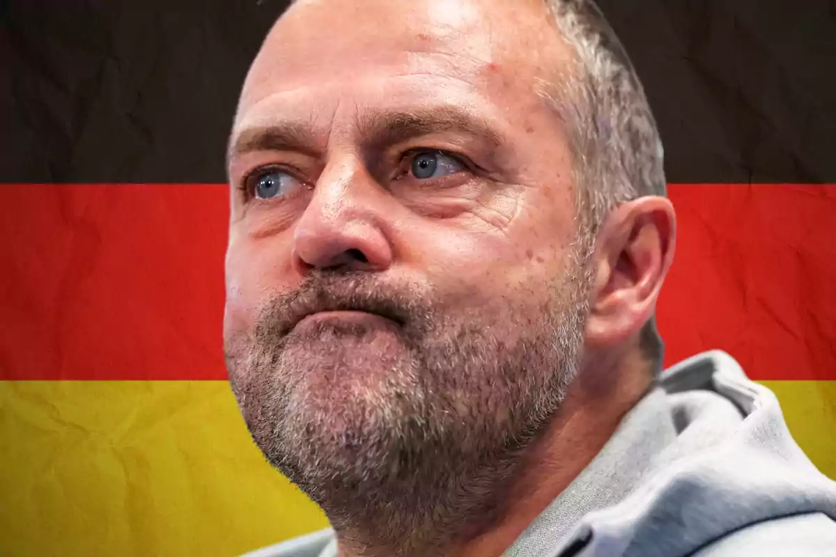 Hombre de expresión seria con barba y cabello corto al frente de una bandera de Alemania desenfocada