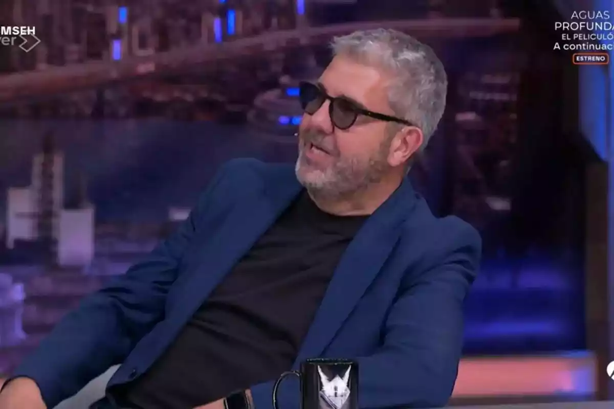 Florentino Fernández con gafas de sol y barba está sentado en El Hormiguero, vistiendo un saco azul y camiseta negra, mientras sonríe y parece estar conversando. Florentino Fernández con gafas de sol y barba está sentado en El Hormiguero, vistiendo un saco azul y camiseta negra, mientras sonríe y parece estar conversando.