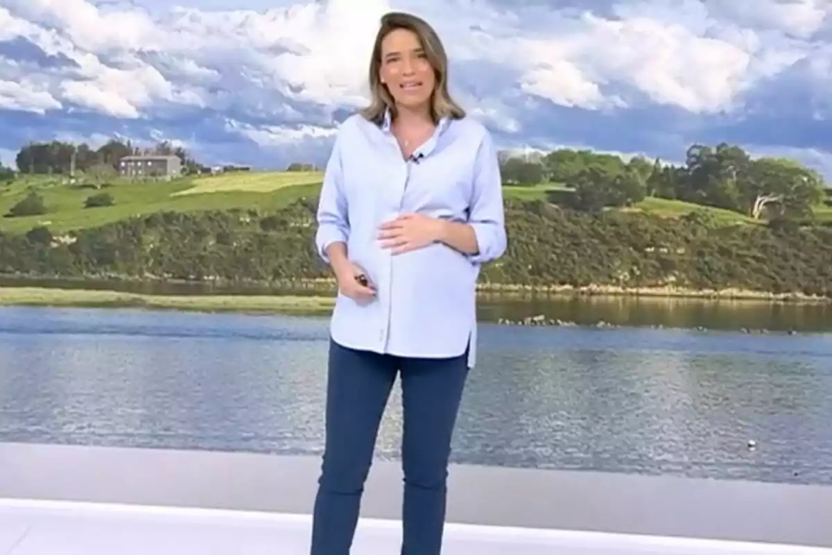 Flora González embarazada está de pie frente a un fondo que muestra un paisaje con un río y colinas verdes bajo un cielo nublado en 'Informativos Telecinco'. Flora González embarazada está de pie frente a un fondo que muestra un paisaje con un río y colinas verdes bajo un cielo nublado en 'Informativos Telecinco'.