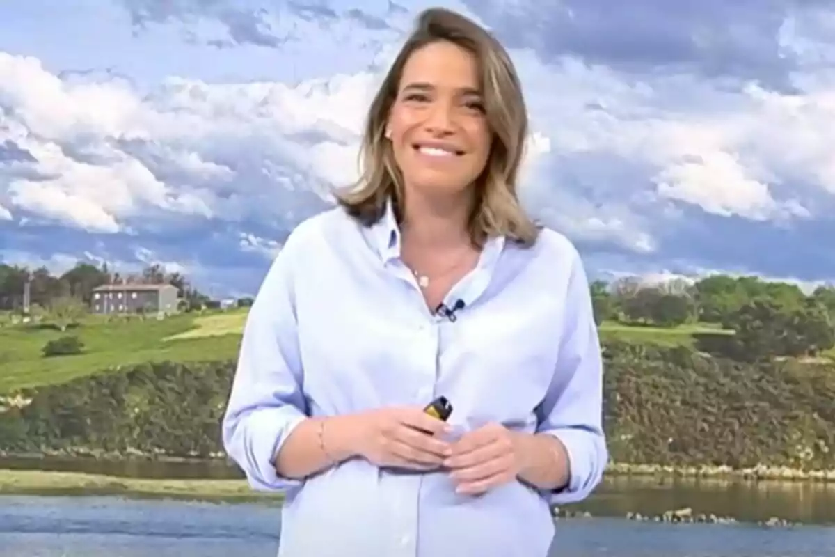 Flora González sonriente con camisa azul claro está de pie frente a un paisaje con cielo nublado y colinas verdes en 'Informativos Telecinco'.