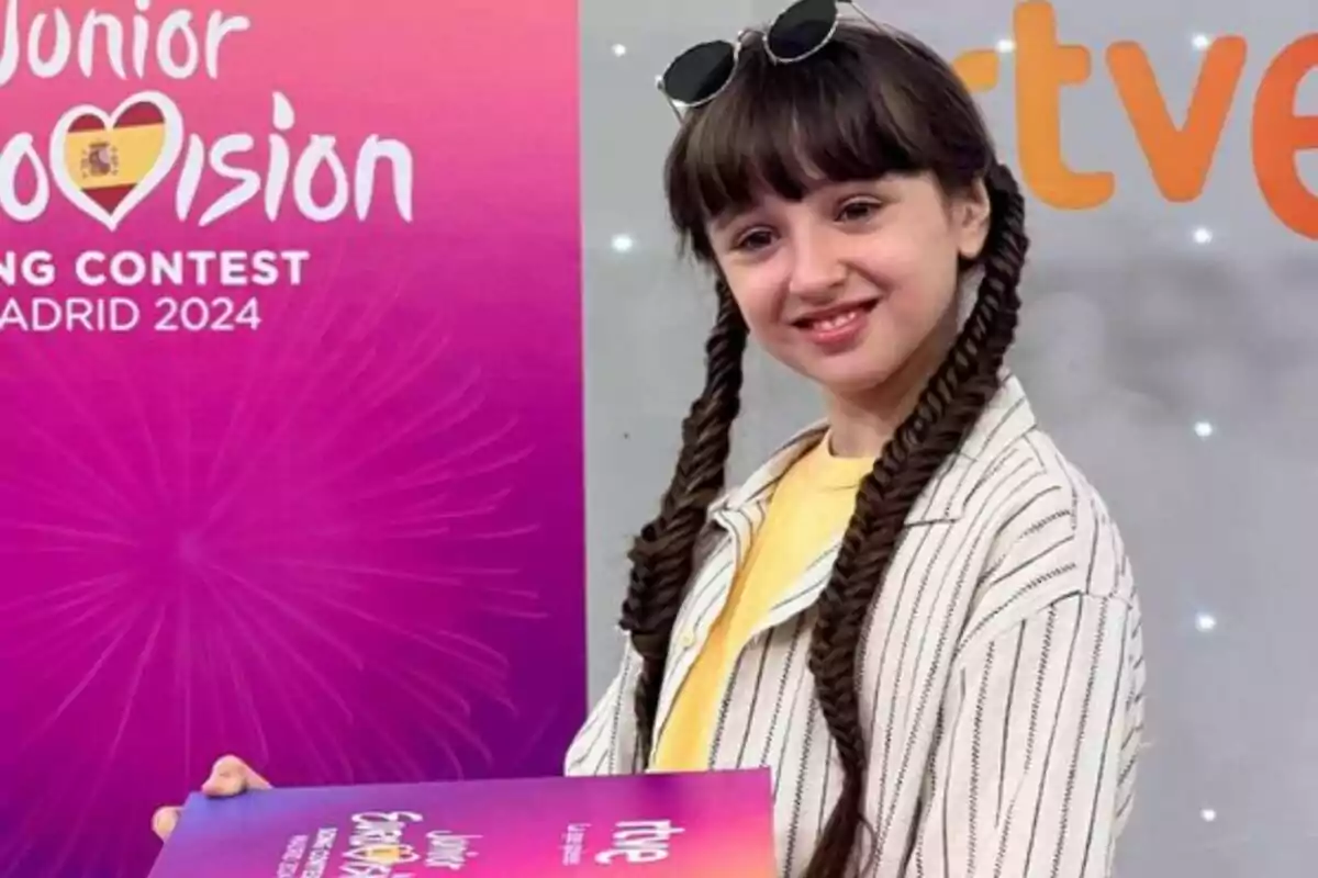 Una niña con trenzas y gafas de sol en la cabeza sostiene un cartel del Junior Eurovision Song Contest Madrid 2024 frente a un fondo rosa con el logo del concurso.