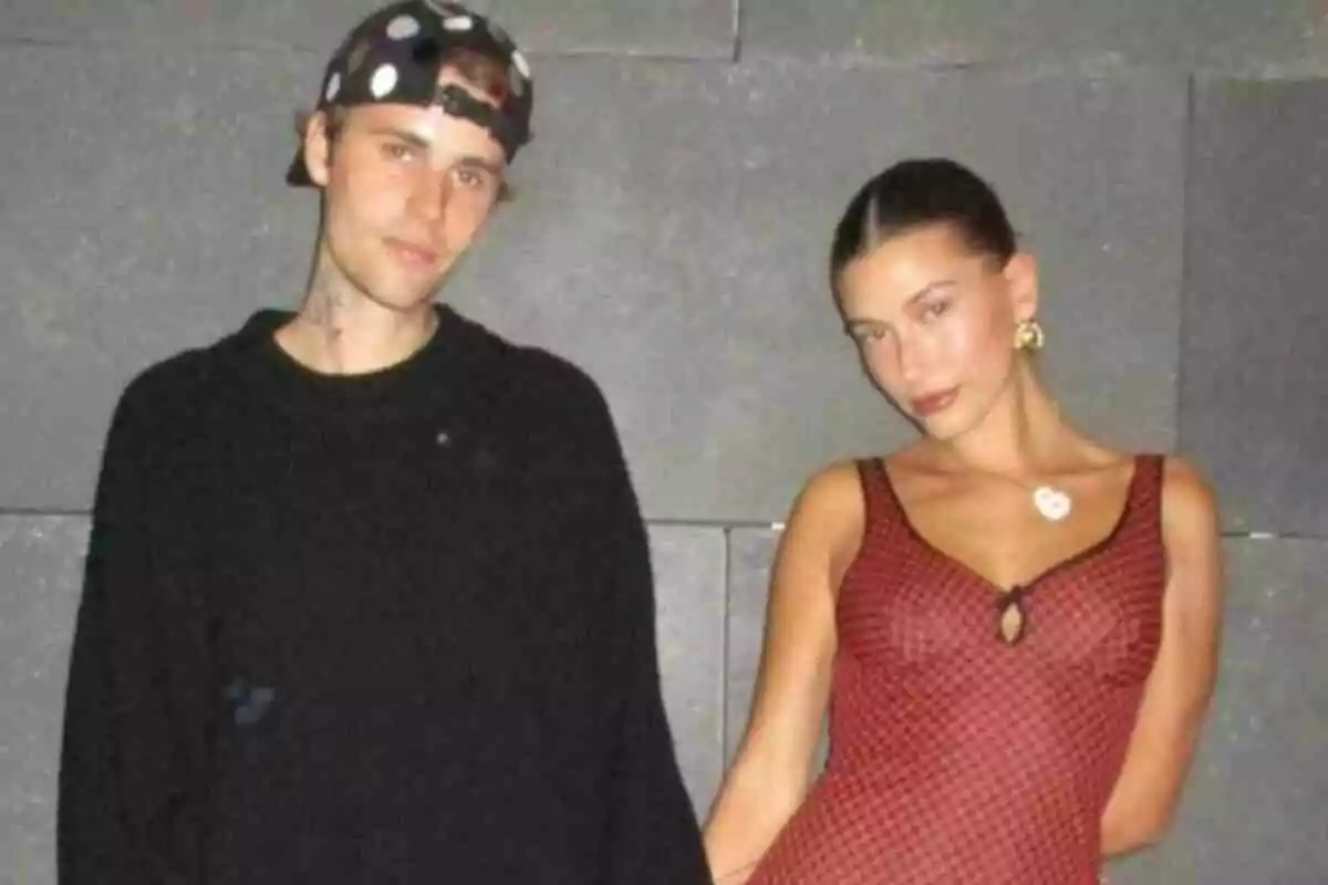 Justin Bieber y Hailey Bieber posando juntas frente a una pared gris, él lleva un suéter negro y gorra, ella un vestido rojo con estampado. Justin Bieber y Hailey Bieber posando juntas frente a una pared gris, él lleva un suéter negro y gorra, ella un vestido rojo con estampado.