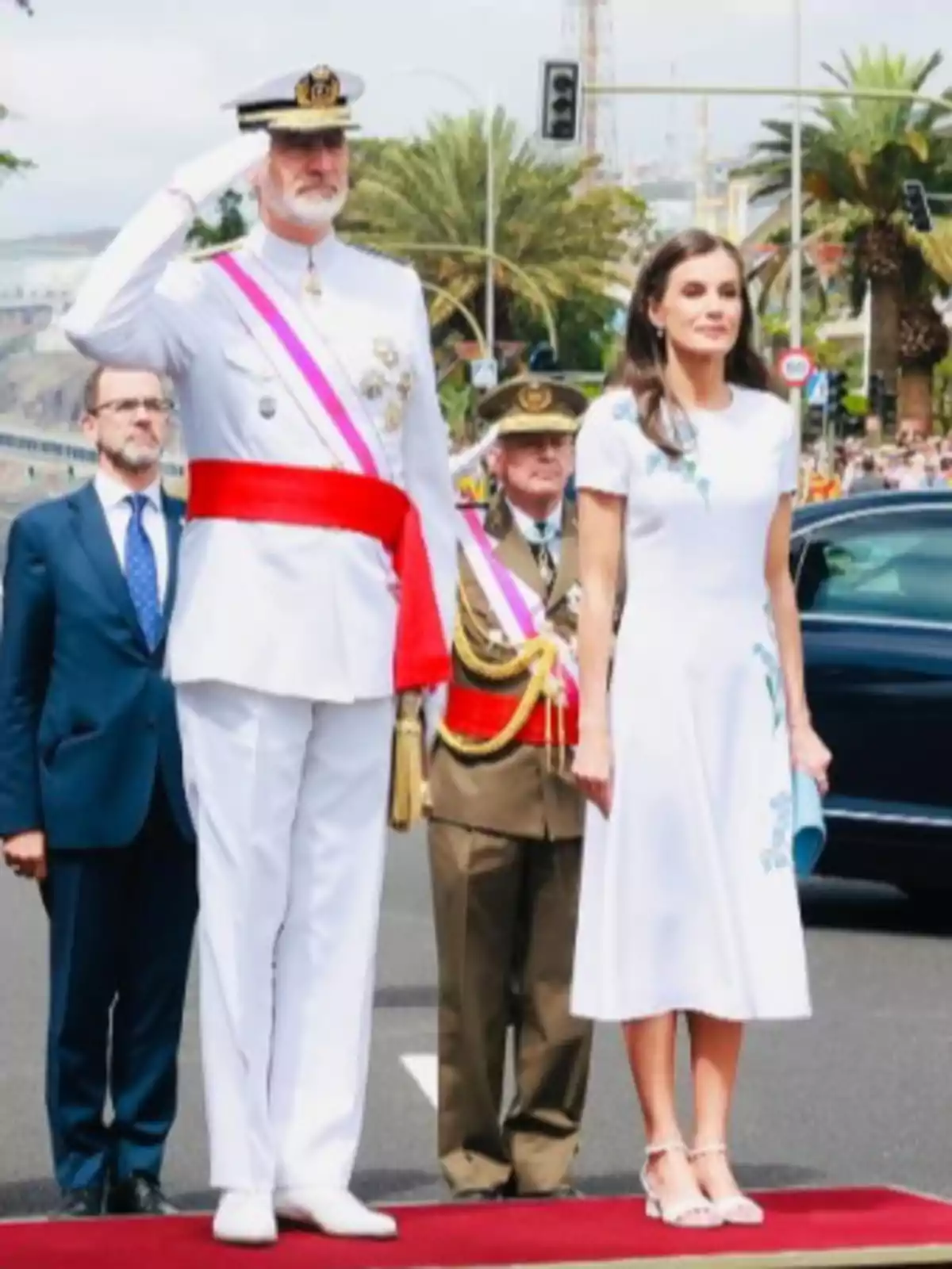 Un hombre con uniforme militar blanco saluda mientras una mujer con vestido blanco está de pie a su lado sobre una alfombra roja en un evento oficial al aire libre Un hombre con uniforme militar blanco saluda mientras una mujer con vestido blanco está de pie a su lado sobre una alfombra roja en un evento oficial al aire libre