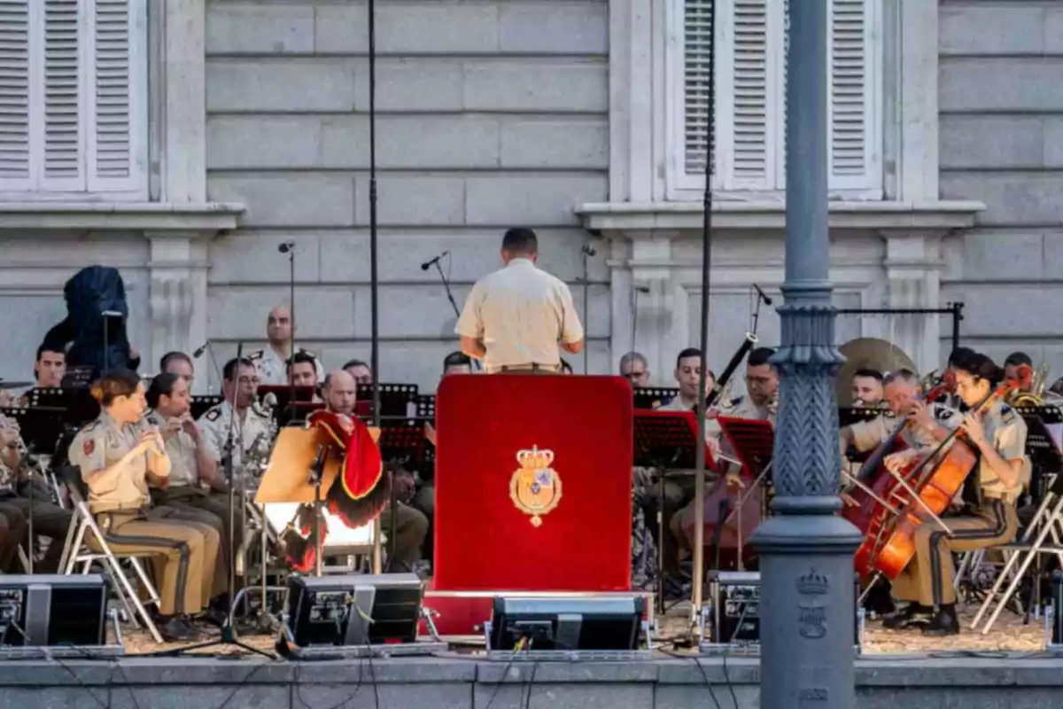 Una banda militar uniformada interpreta música al aire libre bajo la dirección de un director frente a un edificio histórico. Una banda militar uniformada interpreta música al aire libre bajo la dirección de un director frente a un edificio histórico.