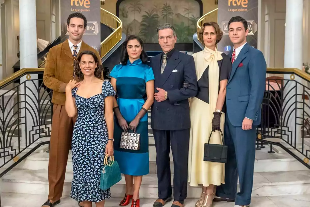 Un grupo de personas elegantemente vestidas posando en una escalera con una decoración de fondo que incluye barandales dorados y un mural en La Moderna