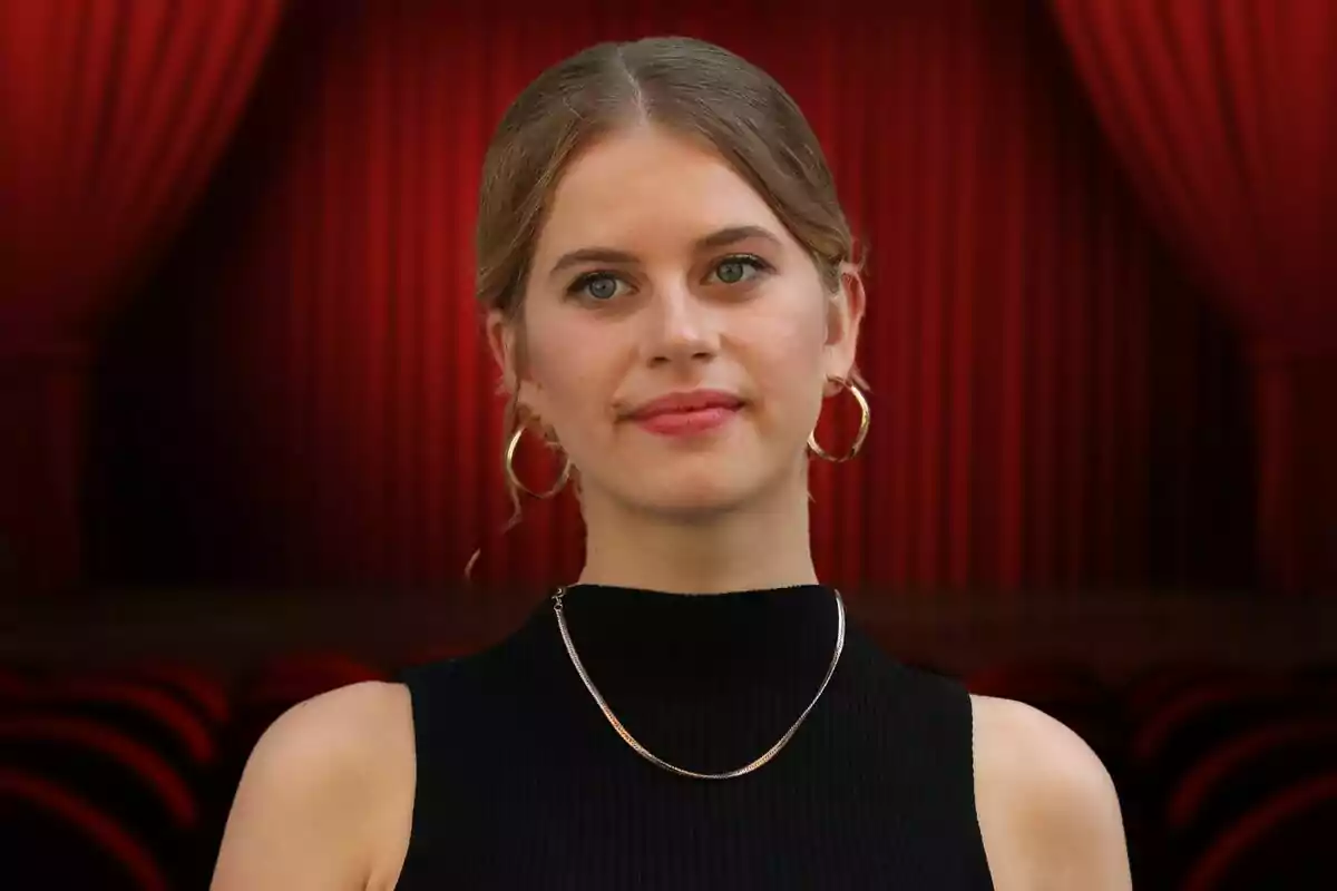 Una mujer joven con cabello rubio recogido, usando un vestido negro sin mangas y aretes grandes, posando frente a un fondo de cortinas rojas de teatro.