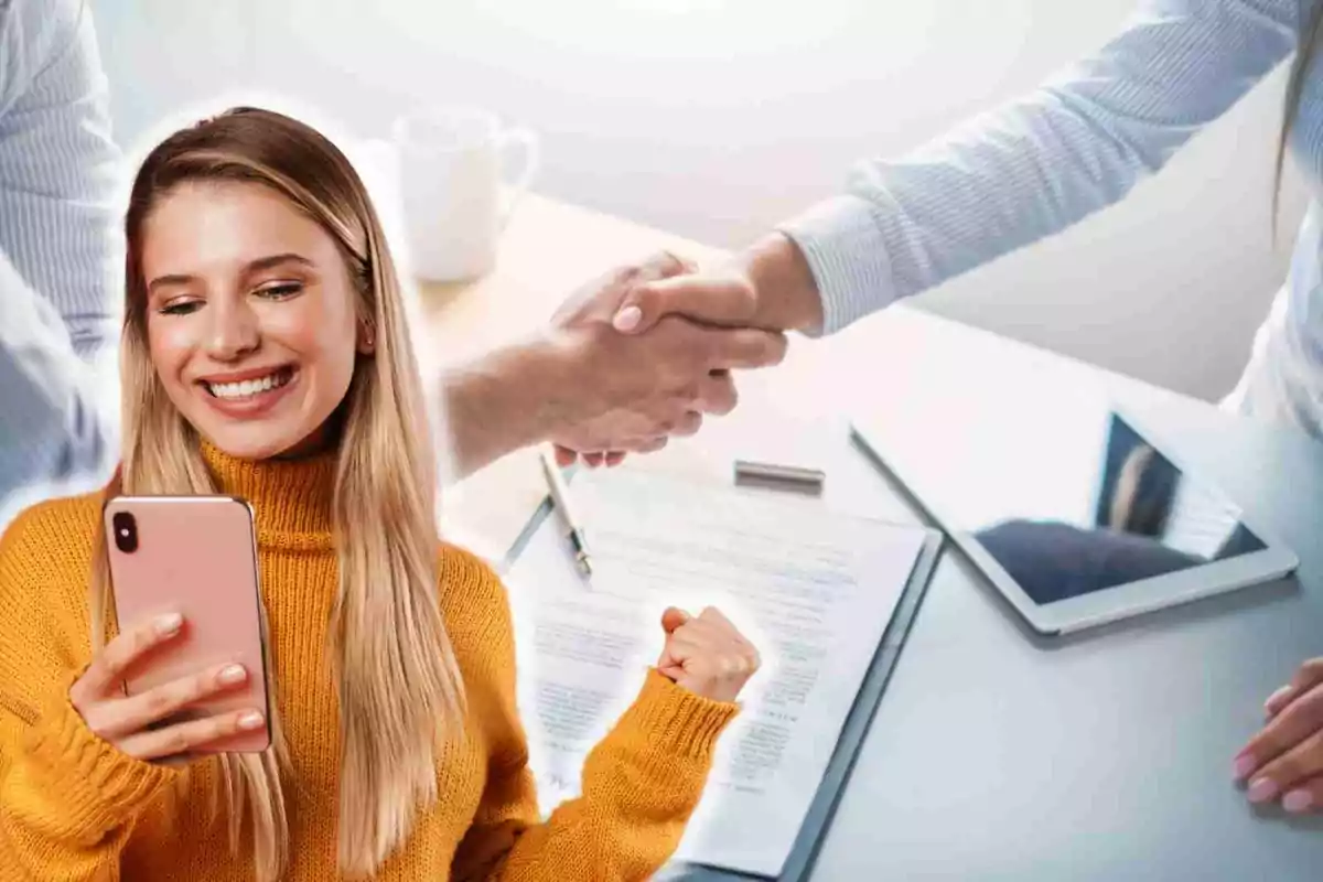 Una mujer sonriente con un suéter amarillo sostiene un teléfono móvil mientras dos personas se dan la mano en el fondo, con documentos y una tableta sobre la mesa.