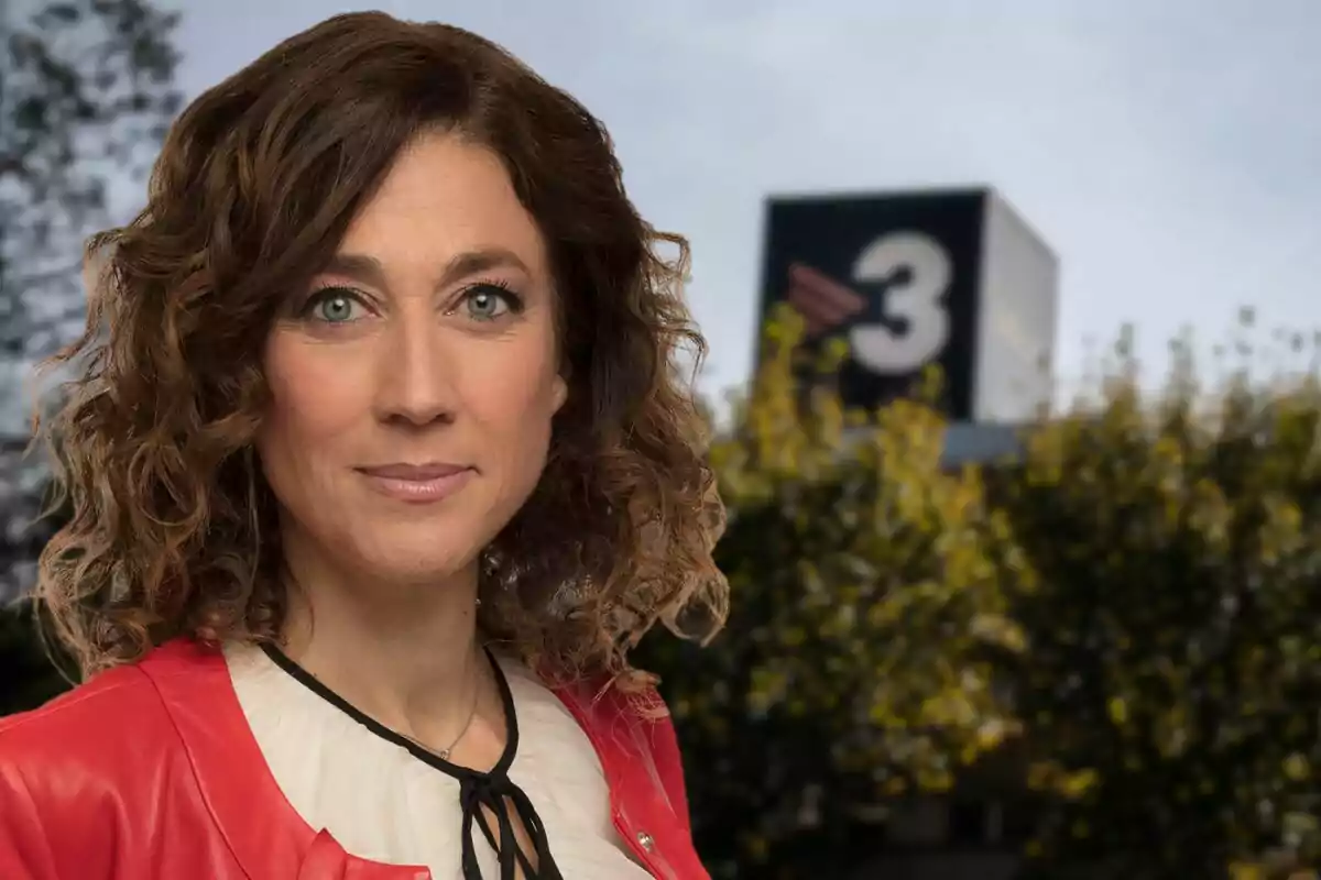 Una mujer con cabello rizado y chaqueta roja está frente a un fondo borroso con un edificio que tiene un número 3.