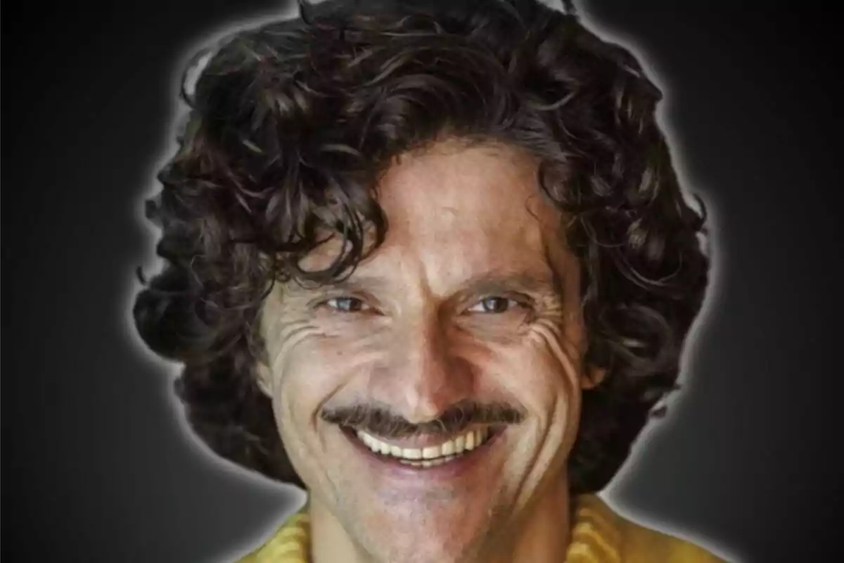 Hombre con cabello rizado y bigote sonriendo frente a un fondo oscuro.