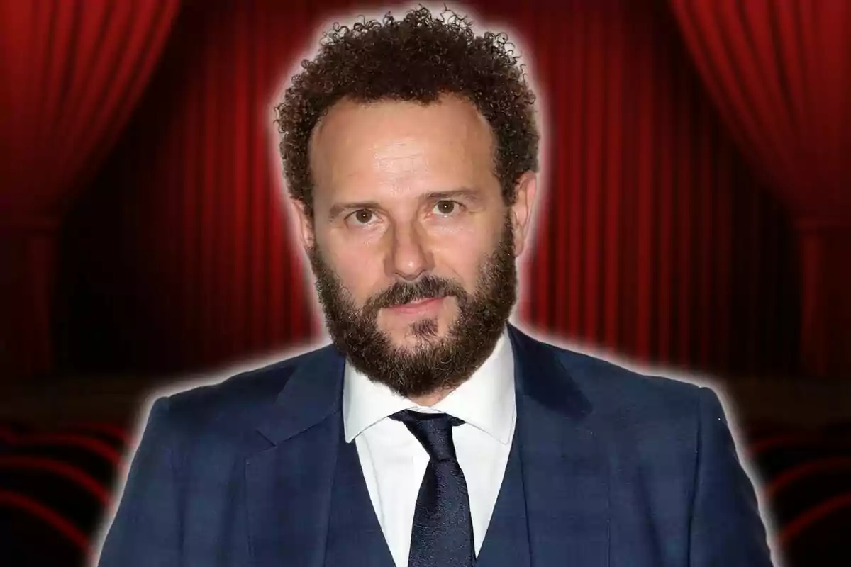 Hombre con barba y cabello rizado, vestido con traje azul y corbata, posando frente a un fondo de cortinas rojas de teatro.