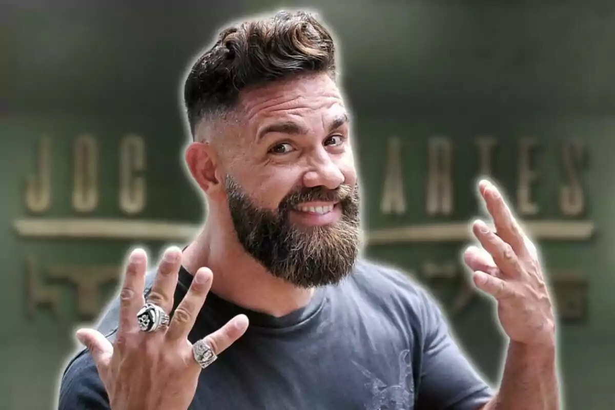 Un hombre con barba y cabello corto sonríe mientras muestra sus manos adornadas con anillos, con un fondo desenfocado que tiene letras.