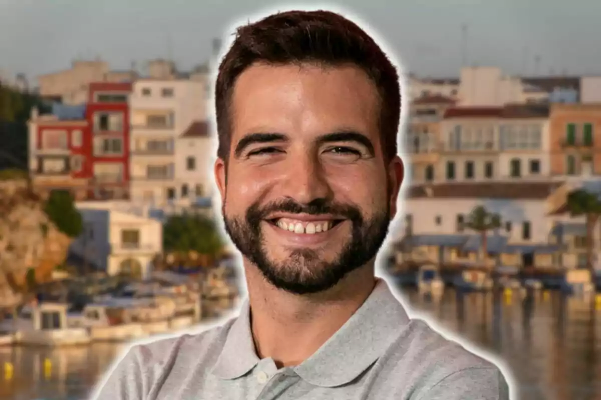 Hombre sonriente con barba y camisa gris frente a un fondo urbano con edificios y un puerto.