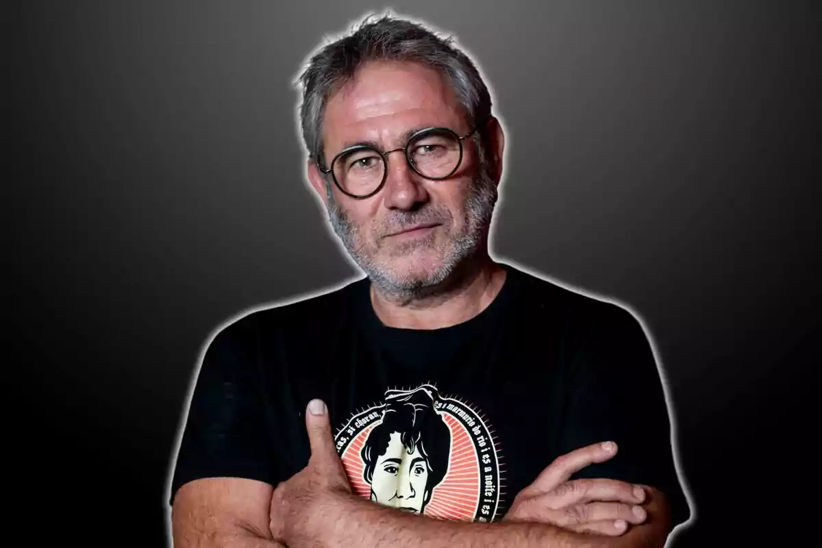 Hombre con gafas y barba gris, con los brazos cruzados, usando una camiseta negra con un diseño gráfico, sobre un fondo gris oscuro.