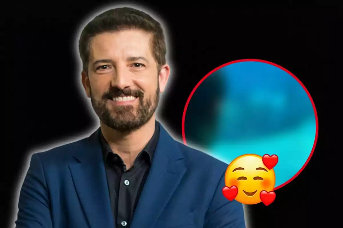 Hombre con barba y traje azul sonriendo frente a un fondo negro con un círculo azul desenfocado y un emoji de cara sonriente con corazones.