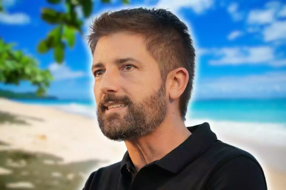 Un hombre con barba y cabello corto mira hacia la derecha mientras está en una playa con el mar y el cielo azul de fondo.