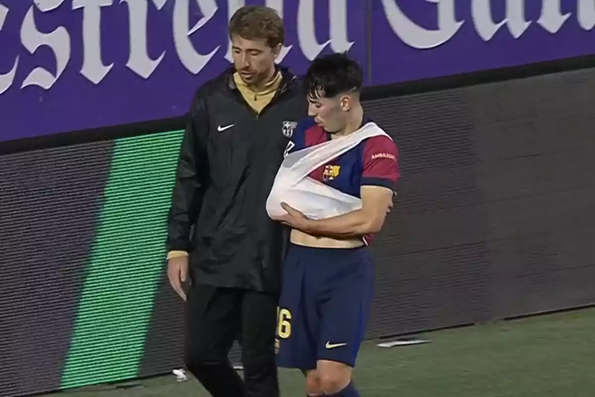 Un jugador de fútbol con el brazo en cabestrillo es acompañado por un miembro del personal en el campo. Un jugador de fútbol con el brazo en cabestrillo es acompañado por un miembro del personal en el campo.