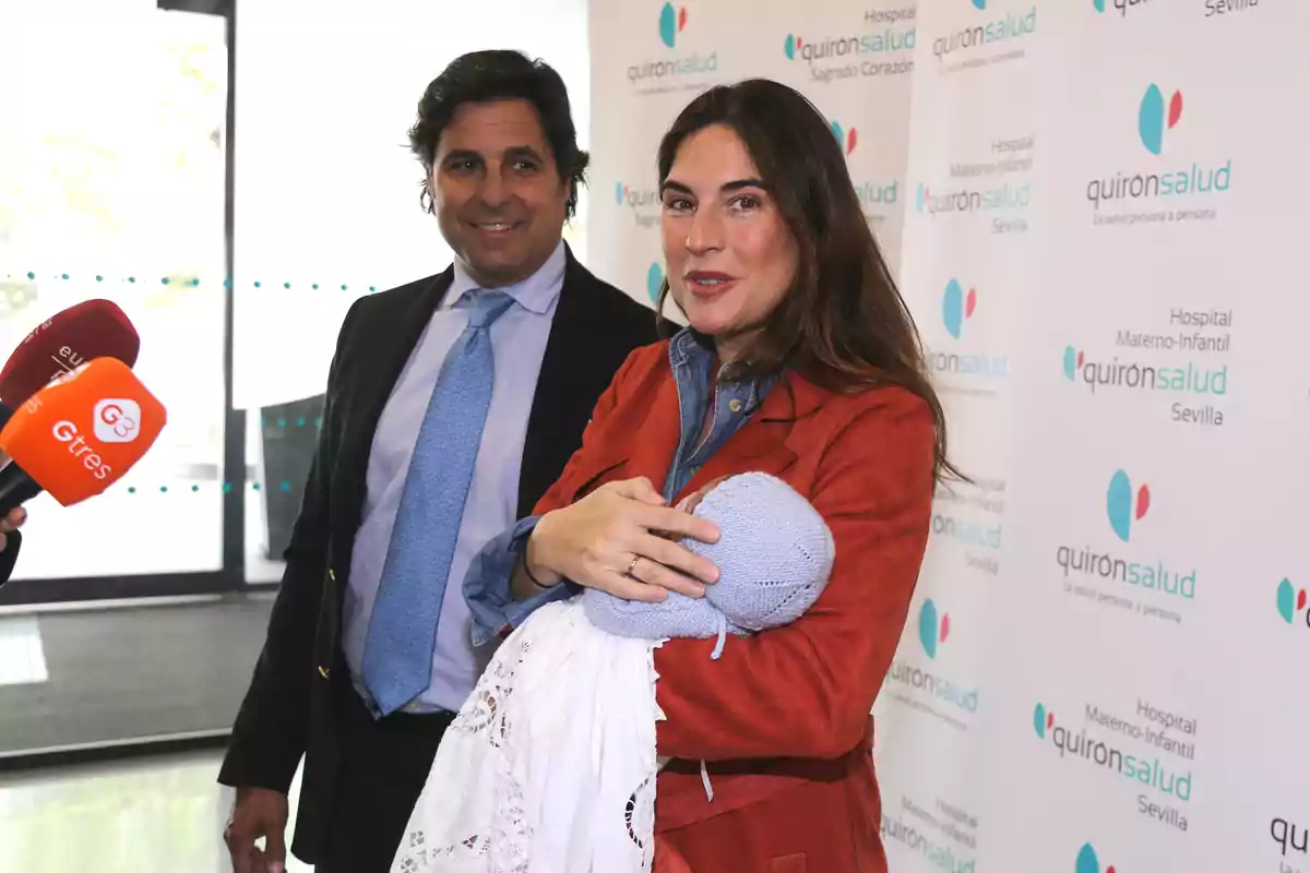 Lourdes Montes sostiene a un bebé envuelto en una manta mientras Fran Rivera sonríe a su lado frente a un fondo con el logo de Quirónsalud y varios micrófonos de prensa. Lourdes Montes sostiene a un bebé envuelto en una manta mientras Fran Rivera sonríe a su lado frente a un fondo con el logo de Quirónsalud y varios micrófonos de prensa.