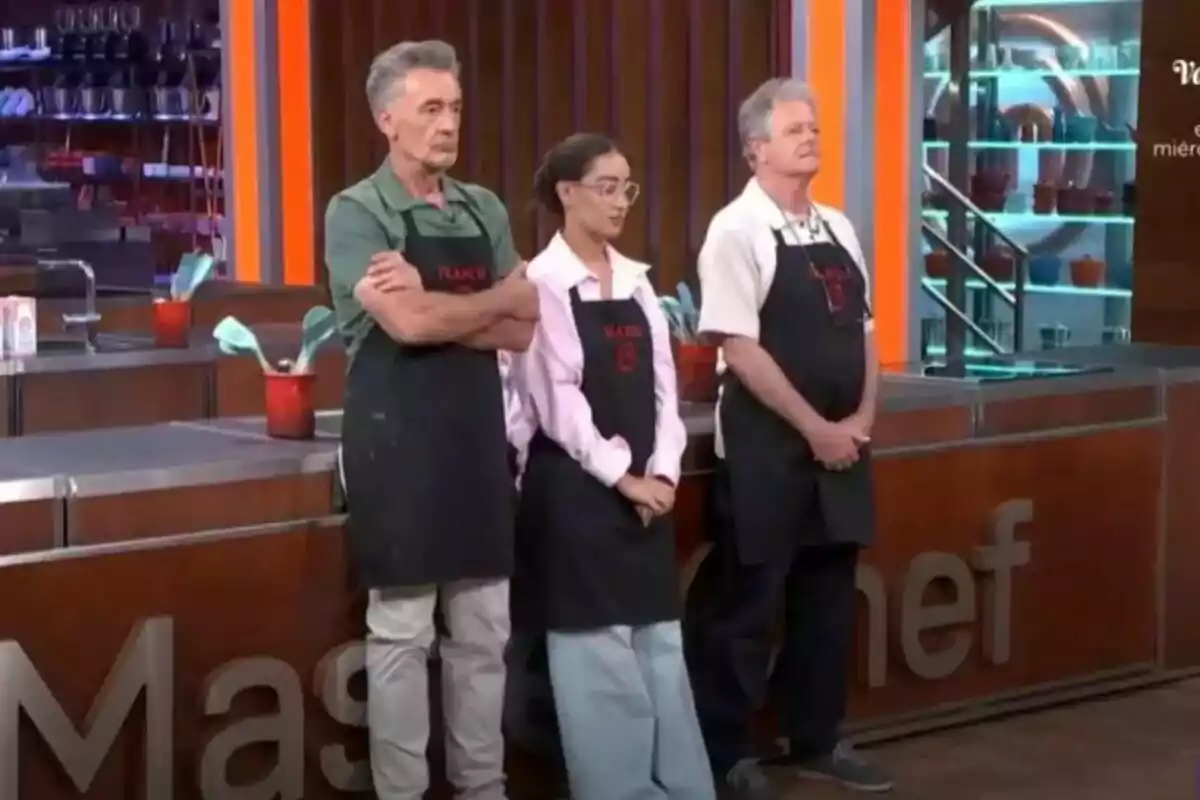 Francis Lorenzo, Marina Rivers y Juan Luis Cano con delantales negros están de pie en una cocina de MasterChef Celebrity.