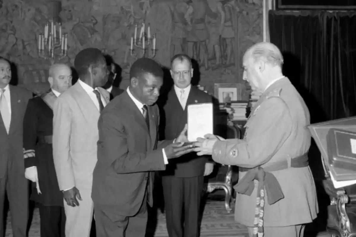 Un hombre de traje recibe un documento de manos de un militar en una ceremonia formal con varias personas observando en un salón elegante Un hombre de traje recibe un documento de manos de un militar en una ceremonia formal con varias personas observando en un salón elegante