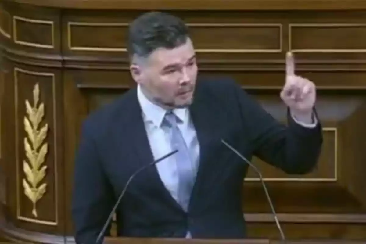 Un hombre de traje y corbata está hablando en un podio de madera, levantando un dedo mientras se dirige al público. Un hombre de traje y corbata está hablando en un podio de madera, levantando un dedo mientras se dirige al público.