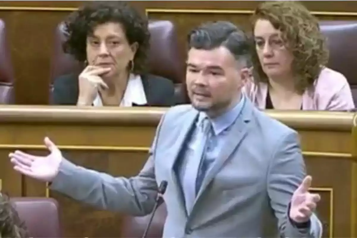 Un hombre de traje gris gesticula mientras habla en un entorno parlamentario, con dos mujeres sentadas detrás de él observando atentamente. Un hombre de traje gris gesticula mientras habla en un entorno parlamentario, con dos mujeres sentadas detrás de él observando atentamente.