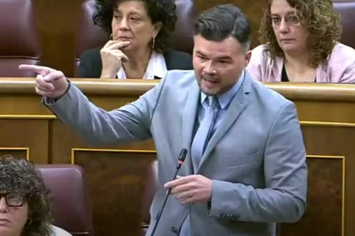 Un hombre de traje gris habla y gesticula en un parlamento mientras otras personas lo observan desde sus asientos.