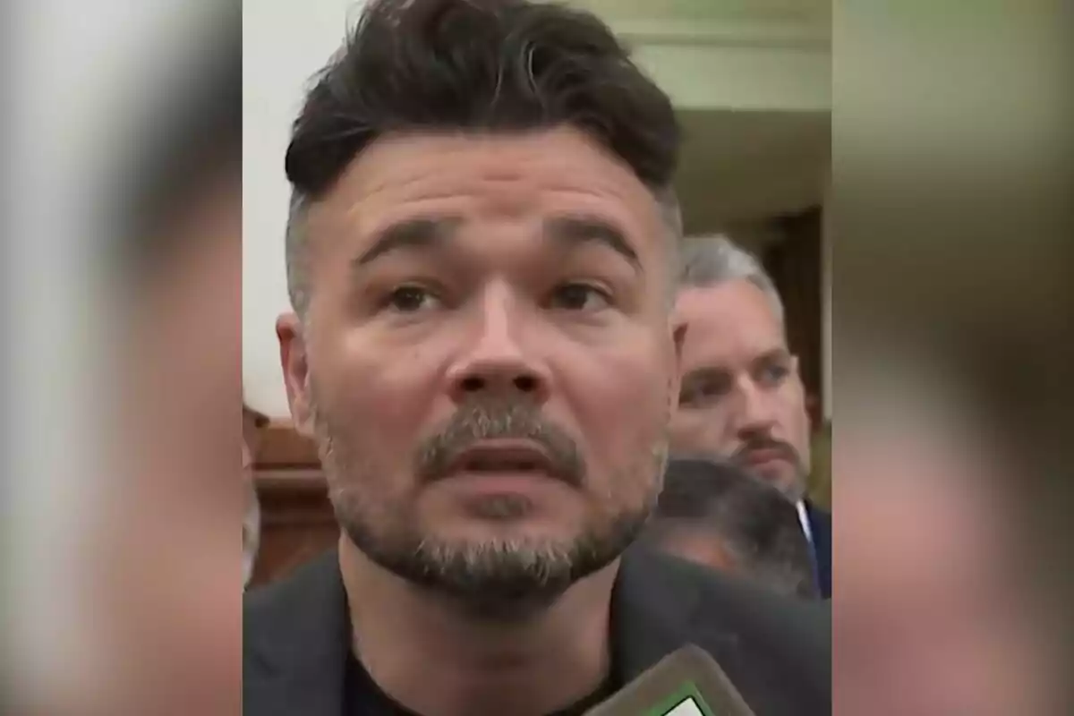 Hombre de cabello oscuro y barba corta hablando frente a un micrófono en un entorno interior con personas al fondo