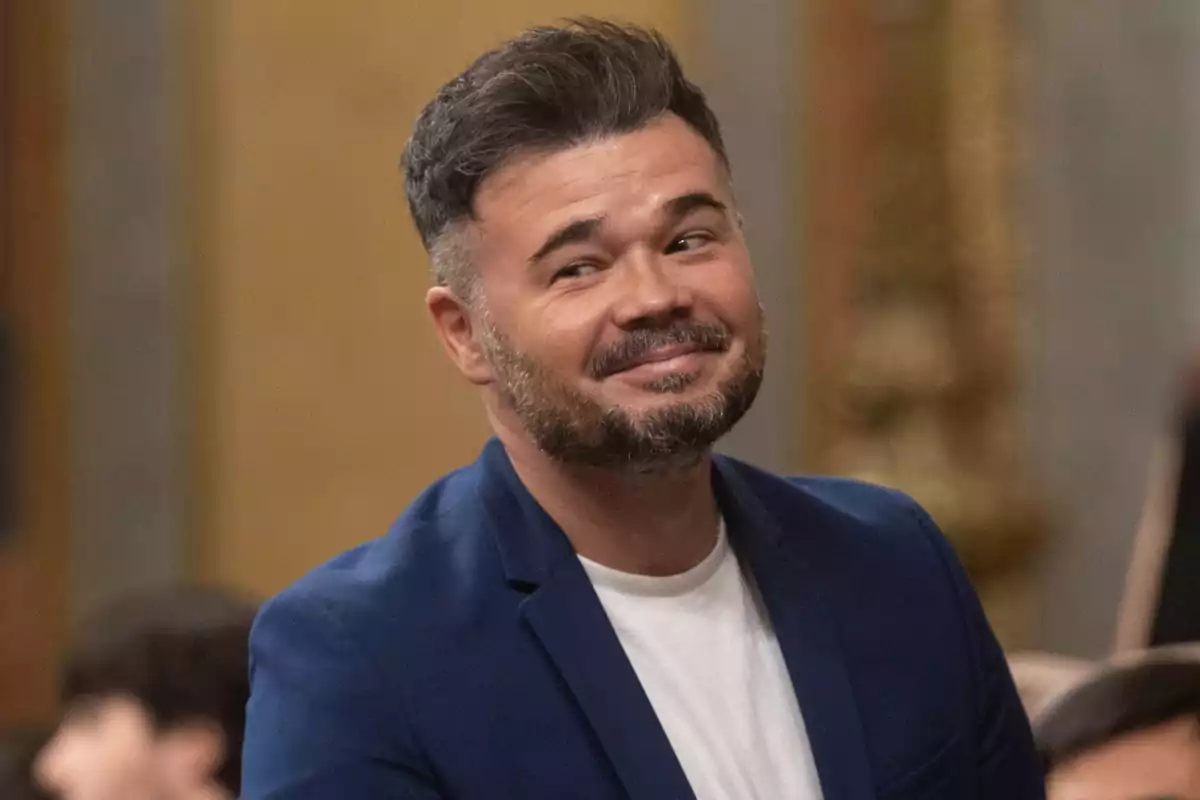 Un hombre con barba y cabello corto, vestido con una camiseta blanca y una chaqueta azul, sonríe mientras mira hacia un lado en un entorno interior.