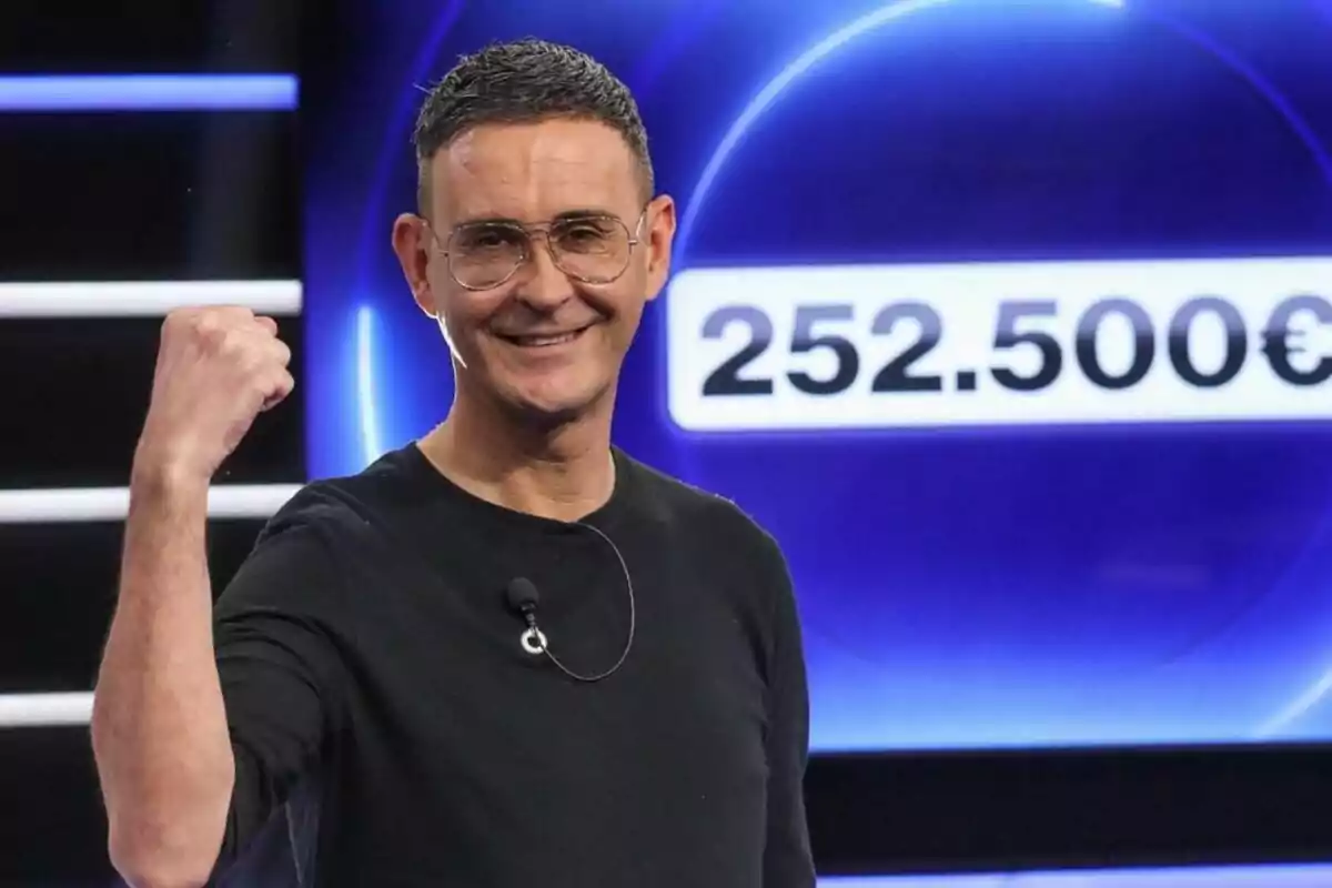 Hombre sonriente con gafas y camiseta negra levantando el puño en señal de victoria frente a un fondo azul con la cifra 252.500 euros de Cifras y Letras Hombre sonriente con gafas y camiseta negra levantando el puño en señal de victoria frente a un fondo azul con la cifra 252.500 euros de Cifras y Letras