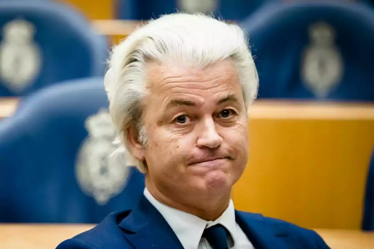 Hombre de cabello canoso y expresión seria sentado en una sala con asientos azules