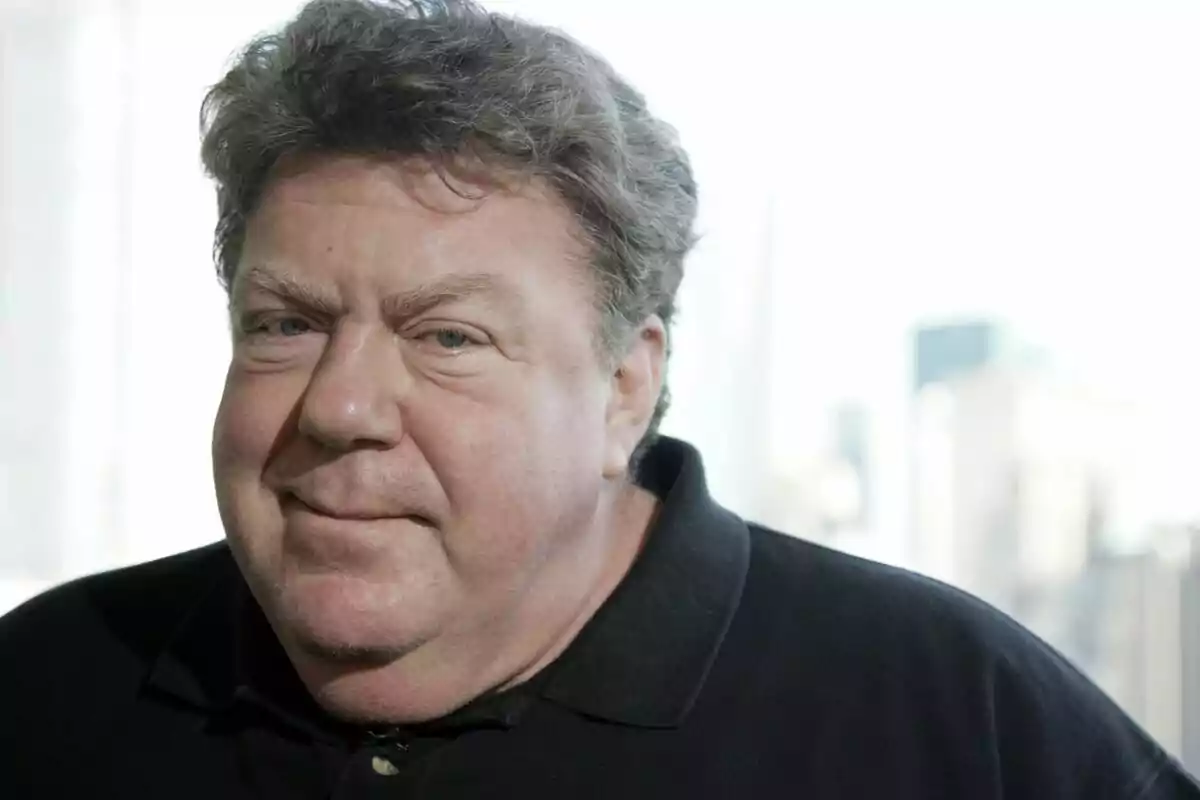 George Wendt con cabello rizado y una expresión neutral lleva una camisa oscura con un fondo desenfocado. George Wendt con cabello rizado y una expresión neutral lleva una camisa oscura con un fondo desenfocado.